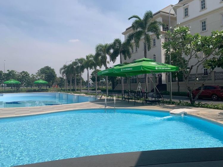 Hồ bơi ngoài trời có hồ nông cho trẻ em, dù che màu xanh lá và hàng cọ, phía sau là dãy nhà nhiều tầng