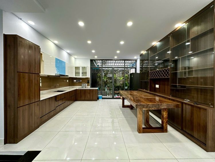 Bếp nhà phố hoàn thiện ở Vạn Phúc City với tủ gỗ tối màu chữ I, bàn ăn gỗ nguyên tấm và cửa kính phía cuối nhà mở ra khoảng sân trồng cây