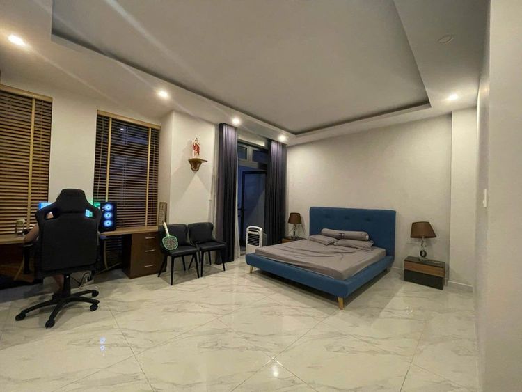Phòng ngủ rộng ở nhà phố 6x17 m có giường bọc nỉ xanh, bàn làm việc và ghế gaming đặt sát cửa sổ, sàn gạch trắng và rèm sẫm màu