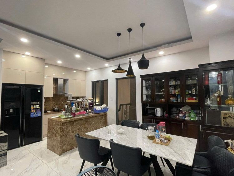 Bếp và phòng ăn nhà phố 6x17 m có đảo bếp ốp đá nâu, bàn ăn mặt đá trắng, tủ lạnh đôi màu đen và tủ rượu kính phía sau