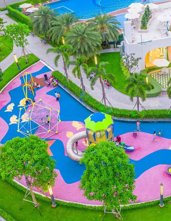 Sân chơi trẻ em lát cao su hồng xanh ở Happy Garden, có lưới leo, cầu trượt ống, cạnh hồ bơi và hàng cọ
