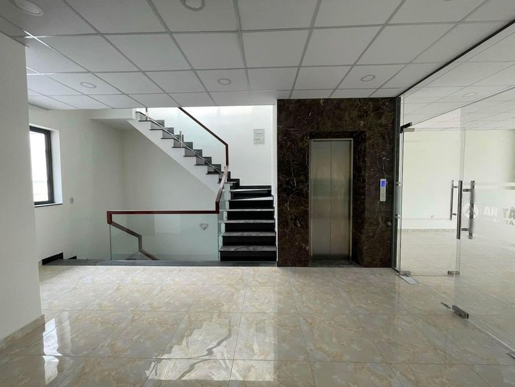 Sảnh trong căn nhà phố 7x20 m hoàn thiện văn phòng với cửa thang máy ốp đá nâu, cầu thang bậc đen tay vịn gỗ và vách kính