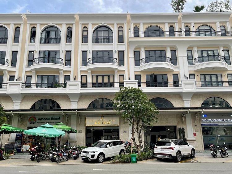 Mặt tiền dãy shophouse 7x20 m tân cổ điển màu kem trên trục Đinh Thị Thi, tầng trệt cửa vòm có biển hiệu, dù xanh và ô tô đỗ trước nhà