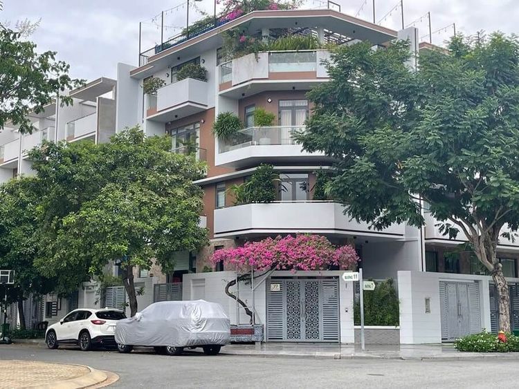 Nhà phố góc 8,6x20 m hai mặt tiền với ban công trồng hoa giấy, cửa cổng trắng và ô tô đỗ trước nhà