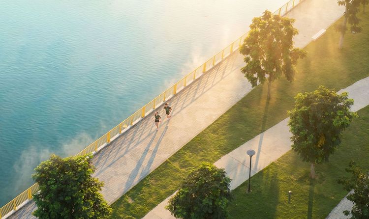Lối đi bộ ven sông có lan can vàng nhìn từ trên cao, nắng sớm chiếu qua hàng cây, hai người đang chạy bộ