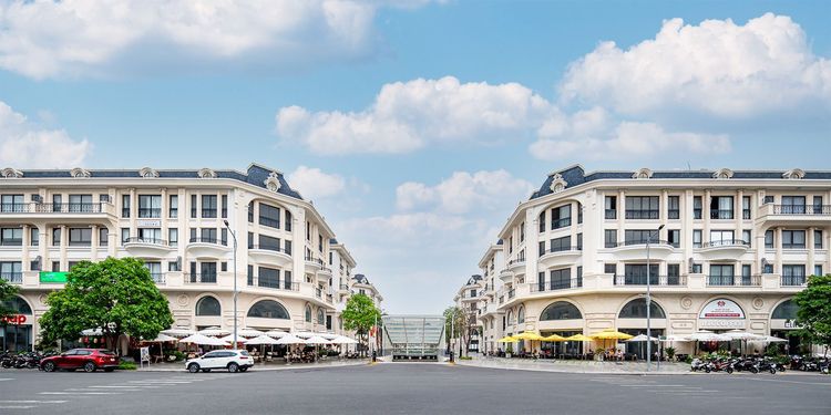 Hai dãy shophouse Royal Vạn Phúc màu trắng kem với mái dốc xám xanh, tầng trệt có dù che quán cà phê, nhìn từ giữa ngã tư