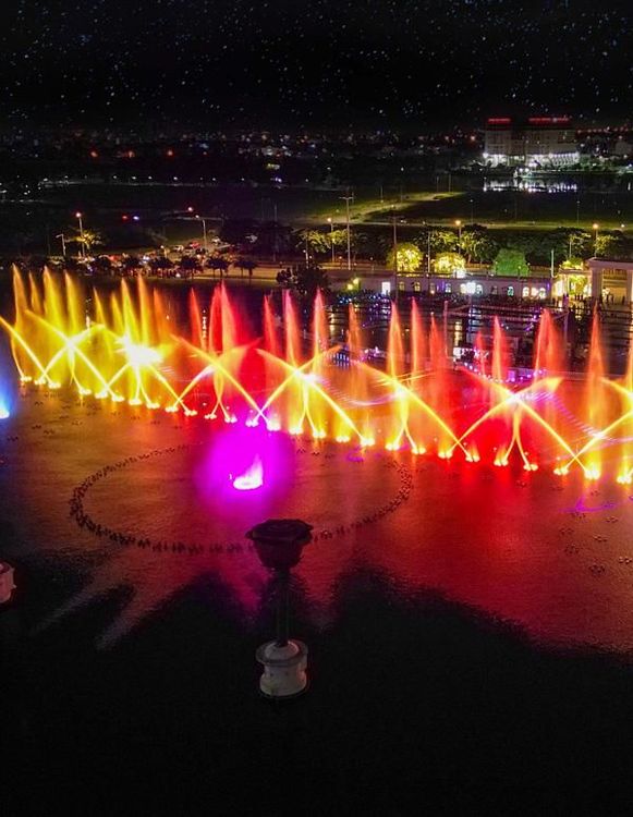 Các cột nước nhiều màu vàng đỏ của show nhạc nước Vạn Phúc City vào ban đêm, phía sau là đèn của khu đô thị