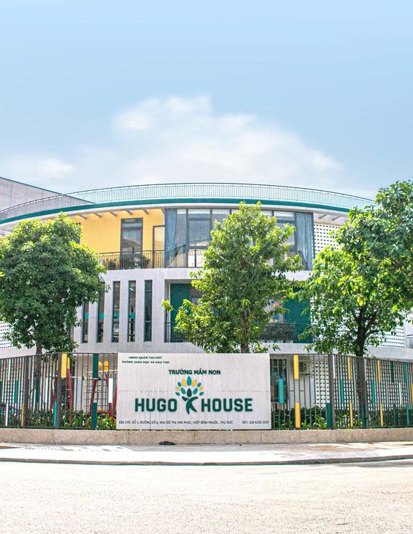 Biển tên Trường mầm non Hugo House đặt trước hàng rào, phía sau là tòa nhà mặt cong nhiều màu và hàng cây